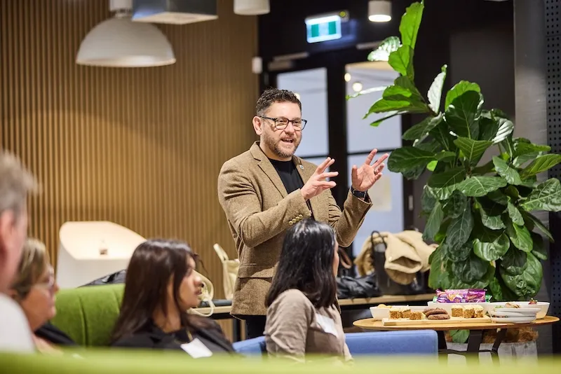 A facilitator in a tweed jacket gestures while speaking to seated Brand Camp participants.