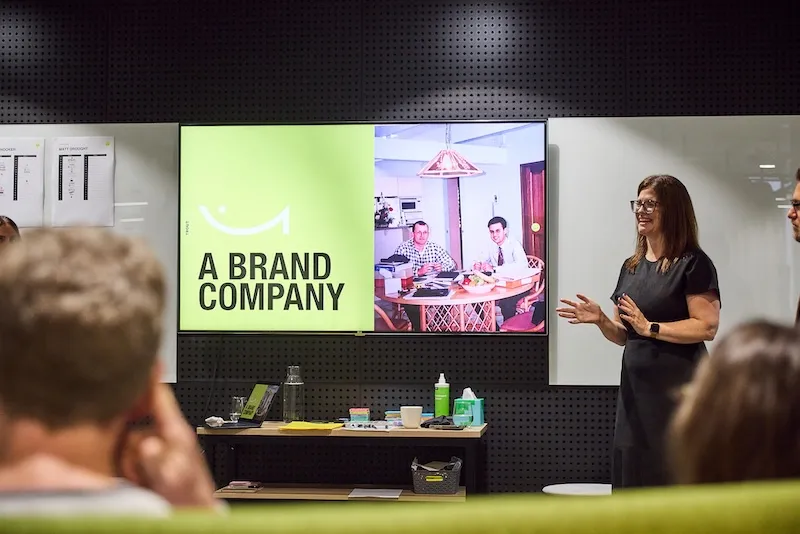 A presenter stands beside a screen reading “A brand company” while the group looks on.
