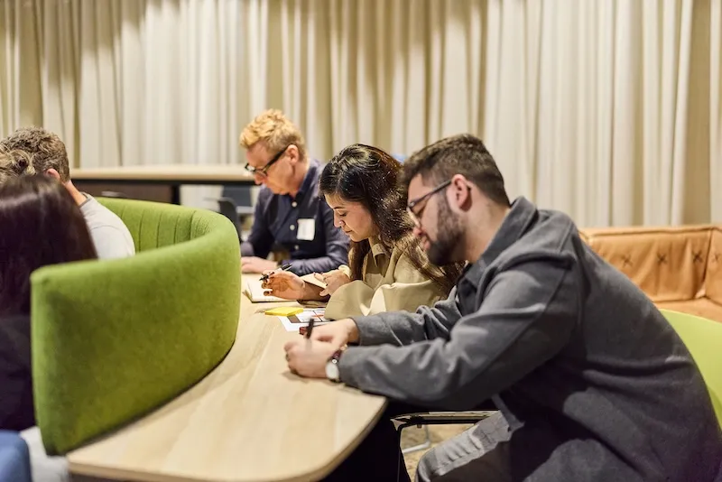 Participants lean over a shared table, writing notes during a workshop exercise.